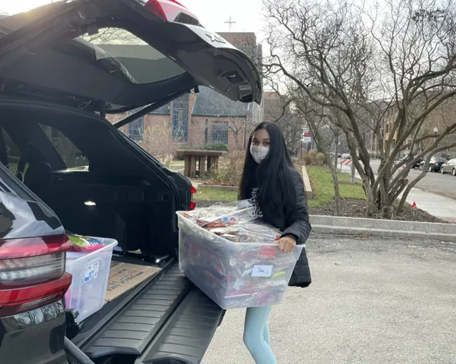 A masked volunteer holding a storage bin beside an open vehicle trunk.