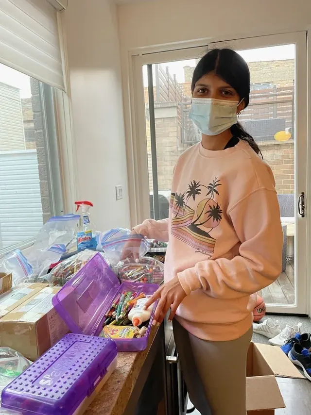 A masked volunteer packing school supplies into bags on a table.