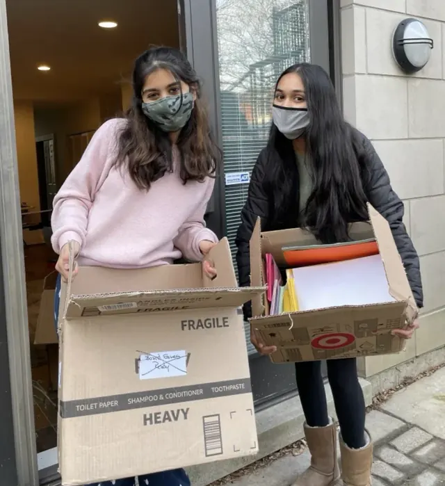 Two masked volunteers carrying open boxes of donated supplies.
