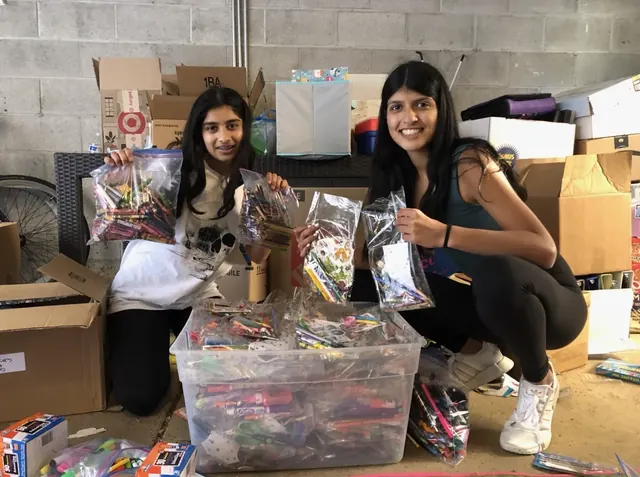 Two volunteers showing completed supply kits beside boxes of donated materials.